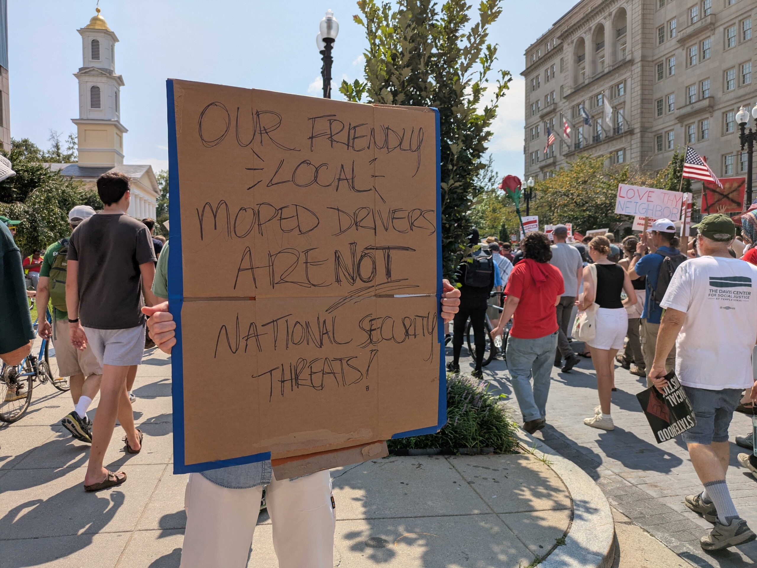 A demonstrator at a march on Sept. 6, 2025, protesting the Trump administration's federalization of law enforcement and deployment of National Guard troops in Washington, D.C., held a sign on 16th Street NW defending local moped food delivery drivers. (Photo by Ashley Murray/States Newsroom)
