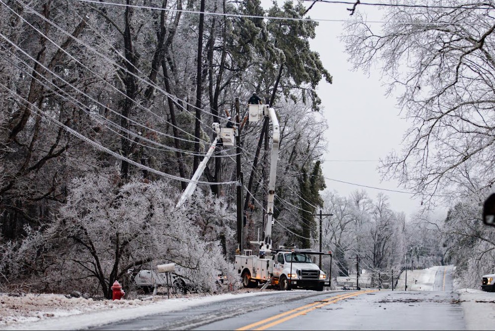 A utility crew working on a tree near power lines in an icy landscape, with an overhead cherry picker and multiple vehicles on a snowy road.